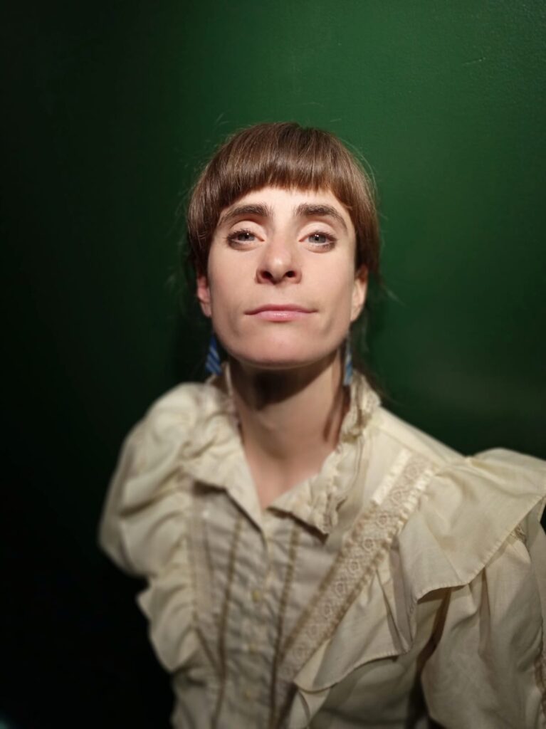 headshot of a woman in a cream coloured long sleeve buttoned shirt against a dark emerald green wall